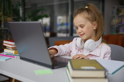 Happy girl child studying online via internet connection using laptop computer in public library while mother reading on blurred background. Portrait of smart little student