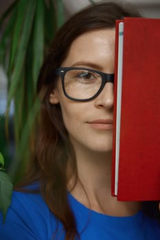 Closeup portrait of teenage girl student covering half part of face with book. Young pretty woman wearing eyeglasses looking at camera with smile