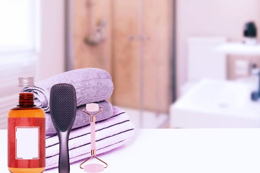 Spa, body, beauty and wellness concept. Various towels, a jade facial roller, a stylish hair brush and a bottle of shampoo over abstract blurred bathroom. Copy space.