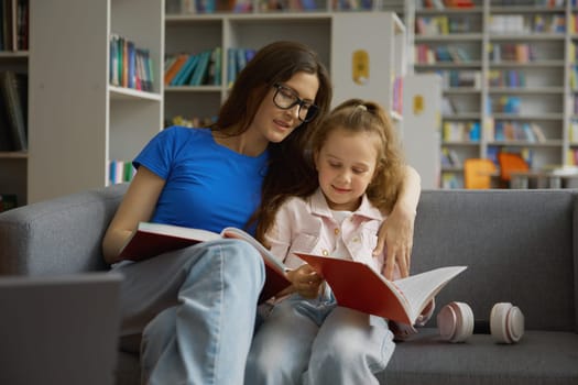 Woman and little girl child taking discussing book for reading while sitting on sofa couch in library or bookstore. Happy family pastime together