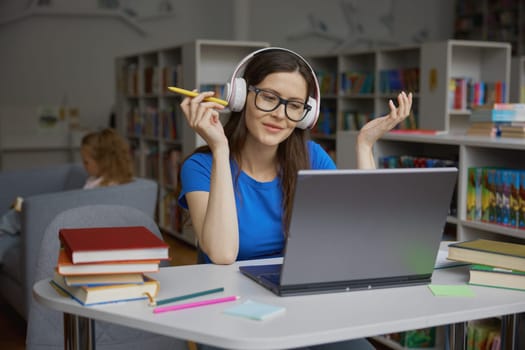 Portrait of smiling positive young woman studying with laptop in library. Teenage student wearing eyeglasses and headphones sitting at table desk looking at camera