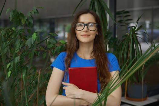 Portrait of young pretty teenage girl student with paper book in hands over green houseplant foliage background. Eco-friendly space for study and learning at library or bookstore concept