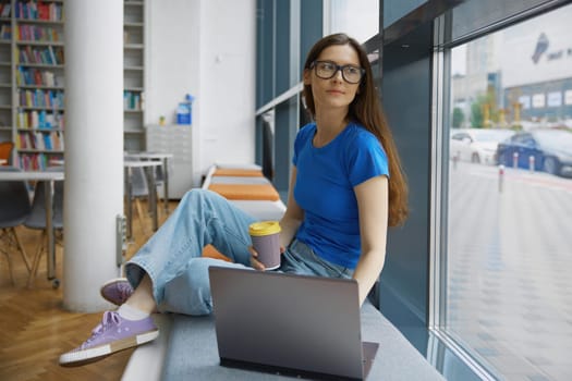 Young woman drinking coffee using laptop for studying in library. Pretty teenage student sitting on windowsill looking at computer screen enjoying aroma beverage browsing in internet at bookstore