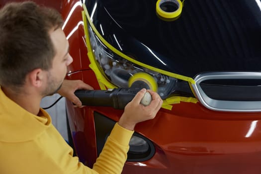 Man technician polishing headlamps using manual power rotary grinder tool with special nozzle while working at car garage workshop