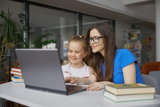 Young woman and girl kid using laptop for studying in library. Tutor and little preschool child reading paper book and online sources on internet