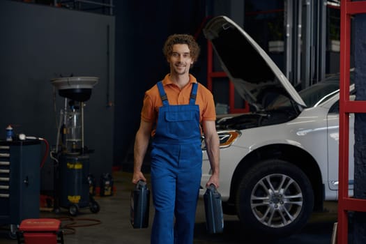 Man auto mechanic wearing overalls carrying canisters with car motor oil walking to camera. Work at automotive service shop