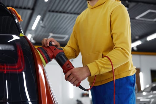 Male auto master providing professional detailing service in car studio. Man mechanic polishing automotive body parts with electric orbital polisher power machine