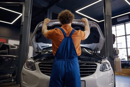 Back view on auto mechanic opening car hood for inspection, diagnostic and troubleshooting of breakage. Professional automobile maintenance service in repair center