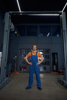 Full length portrait of confident man auto mechanic wearing uniform standing at car lifter over car repair garage interior