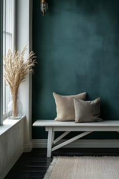 A stylish nook featuring a simple bench, soft cushions, and dried flowers by a large window with a dark green wall.