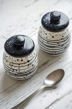 Two decorative ceramic containers with lids sit on a white wood surface alongside a silver spoon, showcasing a modern design.