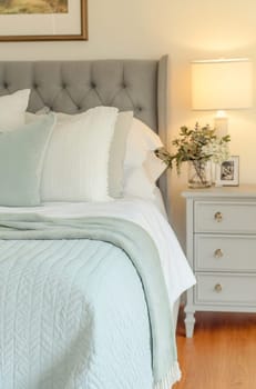 A cozy bedroom features a light gray tufted headboard, soft white bedding, and a bedside table with a lamp and decorative plant.