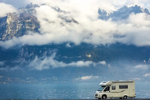 An RV is parked beside a tranquil lake, surrounded by majestic mountains and misty clouds on a peaceful day.