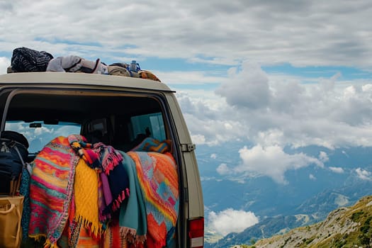 A van is parked on a mountain, displaying colorful blankets on the back, with dramatic clouds and distant peaks visible.
