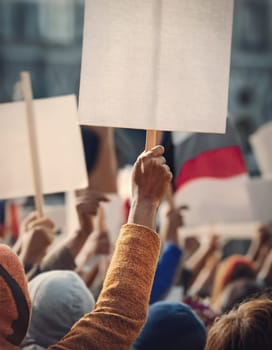 Crowd of people on the street holding up blank protest banners. Hooded man in the foreground holding a placard. Scene of demonstration, riot or rally