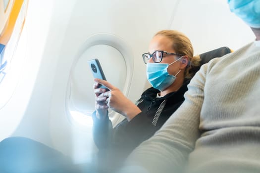 A young woman wearing face mask, using smart phone while traveling on airplane. New normal travel after covid-19 pandemic concept