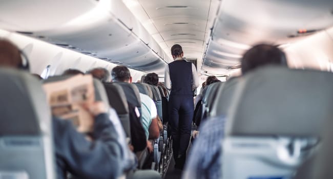 Interior of airplane with passengers on seats and stewardess in uniform walking the aisle, serving people. Commercial economy flight service concept