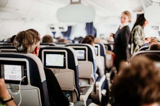 Interior of airplane with passengers on seats and stewardess in uniform walking the aisle, serving people. Commercial economy flight service concept