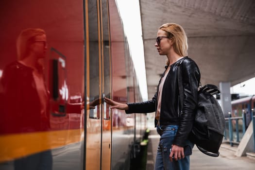 Young blond woman in jeans, shirt and leather jacket wearing bag and sunglass, presses door button of modern speed train to board on train station platform. Travel and transportation.