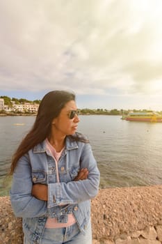 Latina woman in denim jacket and sunglasses looks towards the water under a cloudy sky, porto petro, mallorca balearic islands, spain.