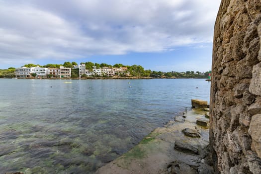 Quiet waterside view of a coastal town with a foreground of stone walls and clouds, porto petro, mallorca balearic islands, spain