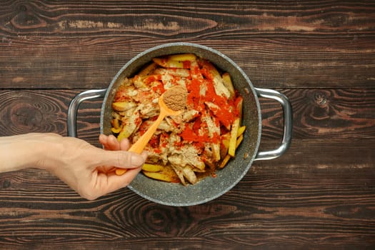 A hand adds coriander to a pot filled with sliced potatoes and bell peppers, preparing a flavorful dish on a rustic wooden table. The vibrant ingredients suggest a hearty, home-cooked meal.