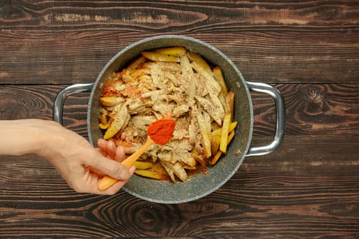 A hand holds a wooden spoon, adding smoked paprika to a pot filled with sliced bell peppers and potatoes. The dish is being prepared on a rustic wooden surface, showcasing a vibrant mix of ingredients.