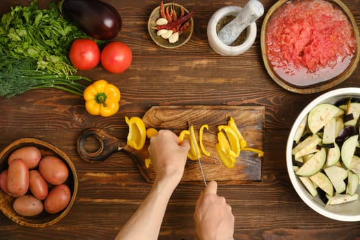 Unrecognizable person chopping bell peppers on a wooden cutting board. Bright yellow peppers, tomatoes, and eggplants are ready for a delicious home-cooked meal.