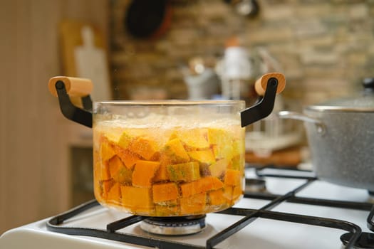 Diced pumpkin simmers in a transparent pot on a gas stove, producing bubbles while the warmth of a cozy kitchen surrounds it. The textured backdrop adds a rustic charm to the cooking process.