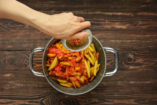 A hand sprinkles chili pepper flakes over a pan filled with fresh tomatoes and potatoes in a warm kitchen. The wooden countertop adds a rustic touch, enhancing the inviting atmosphere of home cooking.
