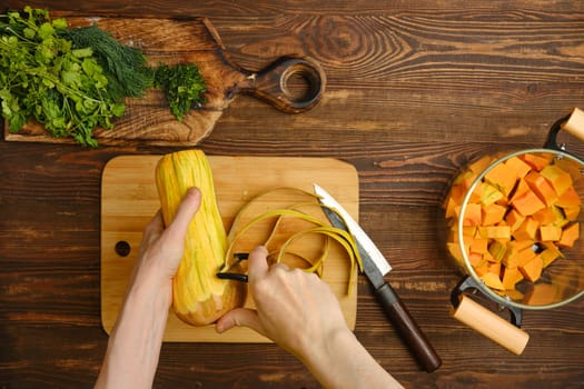 A person is peeling fresh squash with a vegetable peeler, surrounded by freshly chopped herbs and diced squash in a pot, creating a warm and inviting atmosphere in a home kitchen.