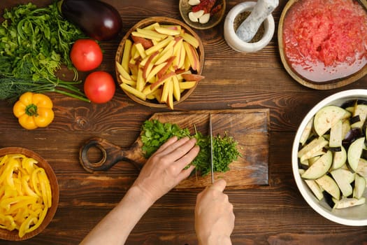 A person chops fresh cilantro on a wooden cutting board while surrounded by various colorful vegetables and green herbs in a warm kitchen. The preparations emphasize a focus on healthy cooking.