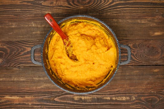 A pot filled with creamy mashed pumpkin on a rustic wooden table. A wooden spoon is partially submerged, showcasing the smooth and vibrant orange texture of the dish.
