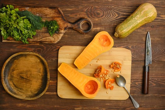 A halved butternut squash on a wooden cutting board alongside fresh herbs. Nearby, a knife and spoon are placed, while a whole squash and a wooden bowl complete the kitchen setting.