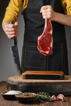 A person in a yellow shirt and apron holds a large cut of raw meat while preparing to slice it on a wooden board. Fresh herbs and spices are arranged nearby, enhancing the culinary experience.