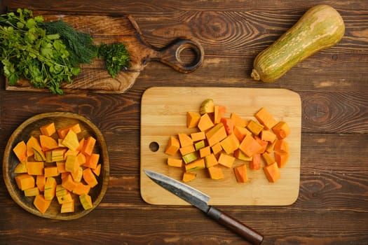 Freshly chopped butternut squash pieces on a wooden cutting board, alongside vibrant herbs like cilantro. A sharp knife rests nearby, ready for meal preparation.
