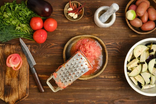 Grated fresh tomatoes and metal grater on a wooden bowl surrounded with eggplant, green herbs and potatoes, are arranged on a wooden table.