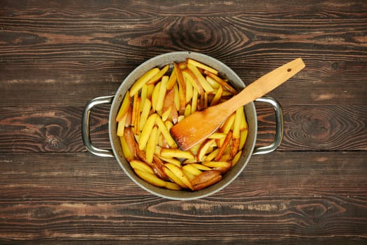 Freshly cut potato fries are frying in a large pot, their golden color starting to develop. A wooden spatula rests inside the pot, ready to stir the crispy fries on a rustic wooden table.