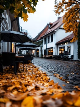 Cozy European cafe street with fallen autumn leaves, cobblestones, and white buildings with warm glowing windows at dusk, outdoor seating area.