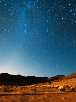 Starry night sky over desert landscape with mountains silhouette and desert brush, illuminated by natural starlight.
