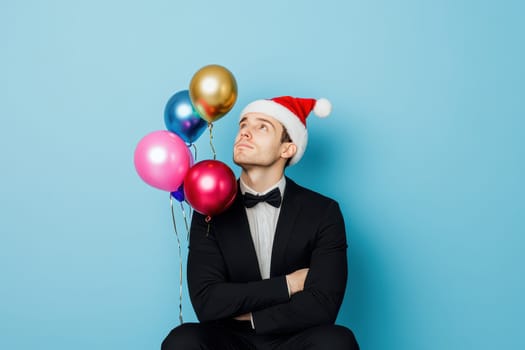 A man wearing a Santa hat sits against a blue background looking up thoughtfully while holding colorful balloons beside him at a celebration.