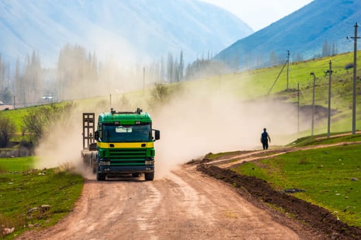 A truck kicks up dust on a dirt road with a pedestrian walking through the haze on a sunny spring day.