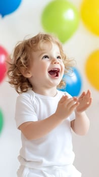 A joyful toddler claps with excitement surrounded by colorful balloons in a bright setting.