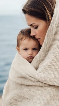 A tender moment between a mother and child wrapped in a blanket by the ocean.