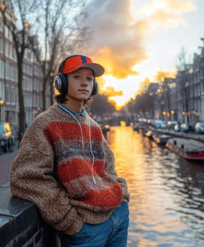 A young man leaning against a wall near the canals, wearing an oversized sweater and a baseball cap, listening to music through his vintage headphones. AI generative.