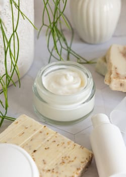 Opened cream jar with white blank lid on white marble against potted green plant and soap bars in bathroom, close up, packaging mockup. Cosmetic product for facecare, beauty routine