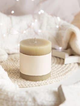 Pillar candle with blank label near white knitted sweater, wooden Christmas decor and lights, copy space. Winter atmospheric mock up in rustic or scandinavian style