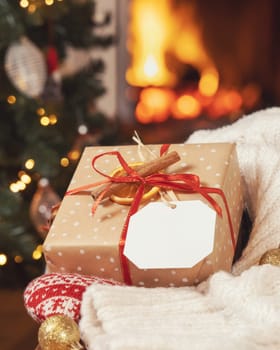 Gift tag on a Christmas present near cosy heart and sweater against Christmas tree lights and burning fireplace, close up, copy space. Warm winter atmospheric mock up, family holiday time
