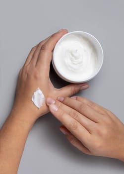 Close up of woman applying protective cream on hand and holding a cosmetic jar in other hand, topview over grey. Beauty treatment or handcare procedure in a spa salon