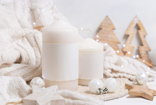 Pillar candle with blank label near white knitted sweater, wooden Christmas decor and lights, copy space. Winter atmospheric mock up in rustic or scandinavian style